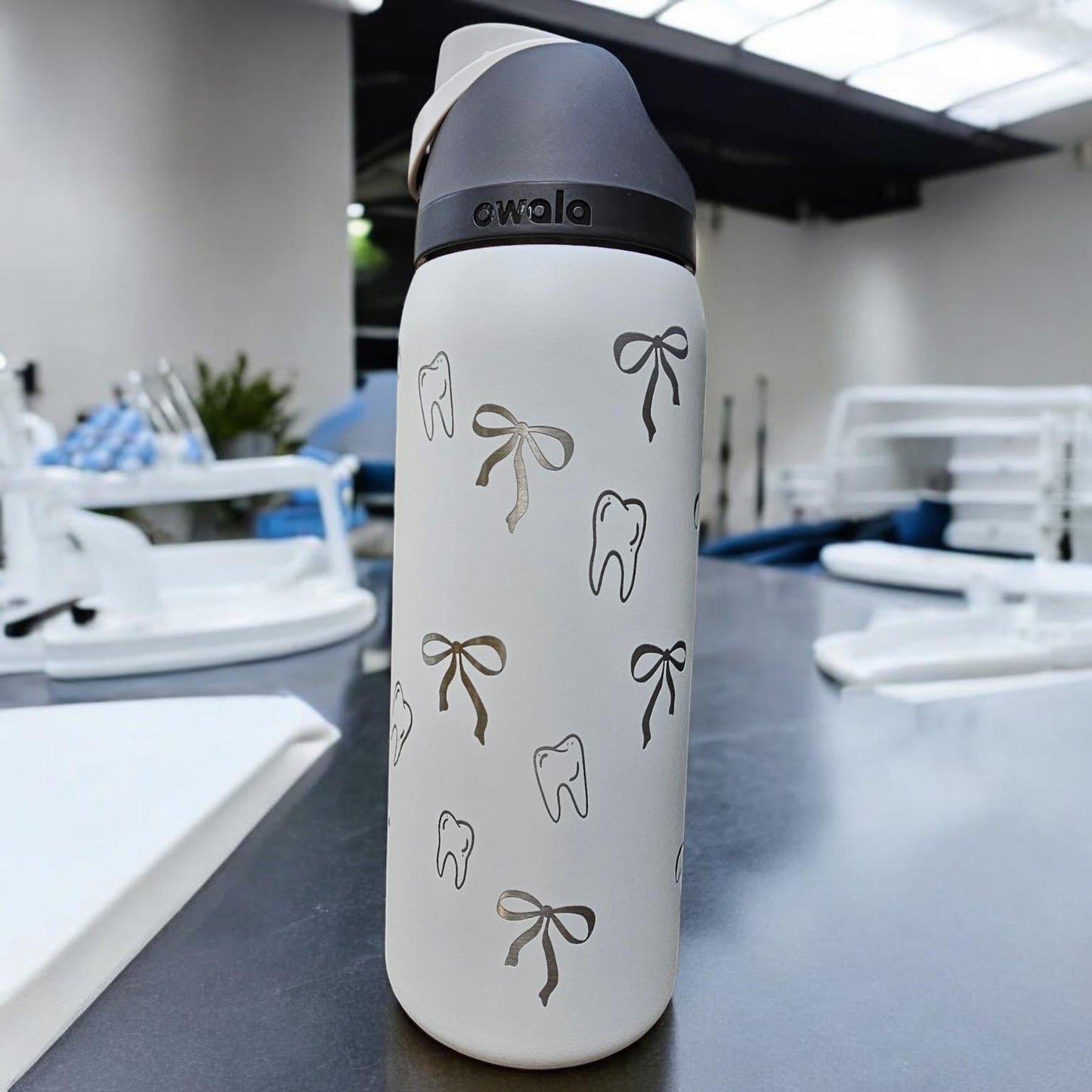 Sleek or Shy Marshmallow Owala water bottle. The bottle features a repeating pattern of small, engraved teeth and bows. The brand name 'Owala' is subtly engraved on the bottle's body. The bottle is shown on a dark gray surface.