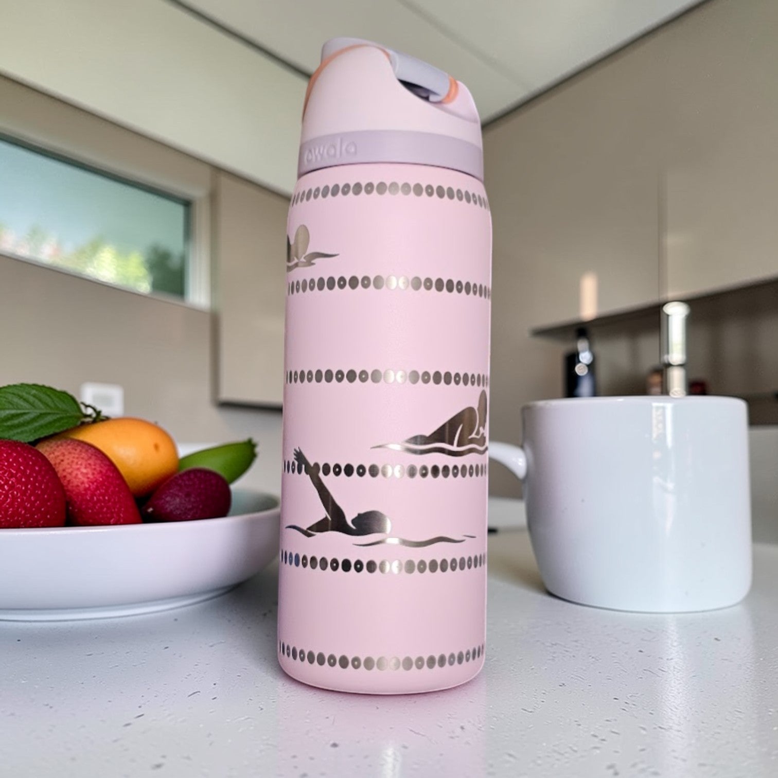 Pink thermos bottle with animal design on a kitchen counter with fruit and a mug.