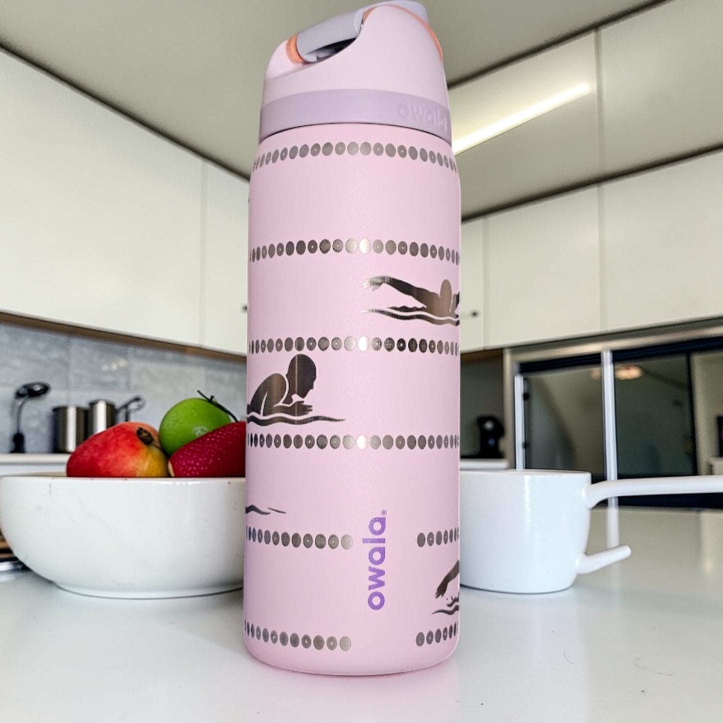Pink Owala water bottle on a kitchen counter with fruits and a bowl in the background